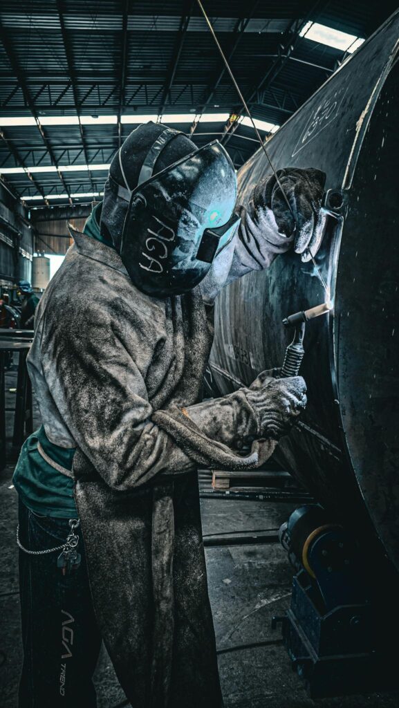 A welder in protective gear welding metal structures in a factory environment.